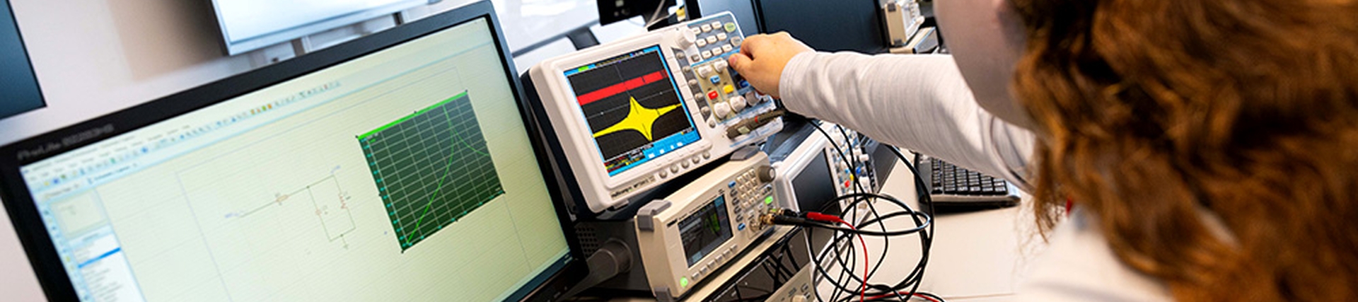A young woman with long red hair adjusts a setting on a device; the monitor in front of her displays a line graph.