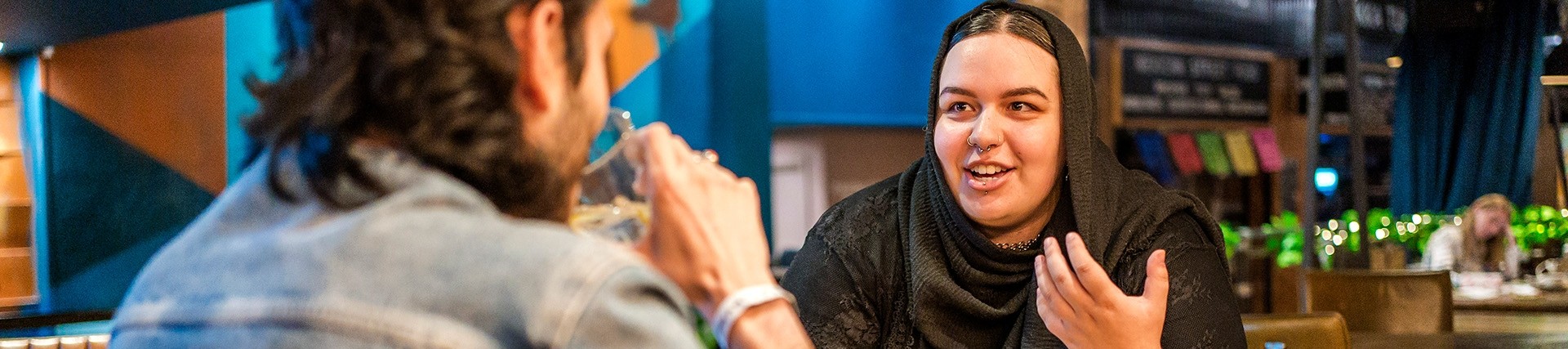 A young woman wearing a hijab chats to a friend in a modern café-bar.