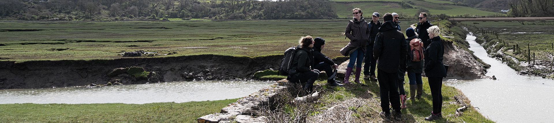 A group of students in outdoor gear stand on a turf bank above the Cwm Ivy salt marsh.