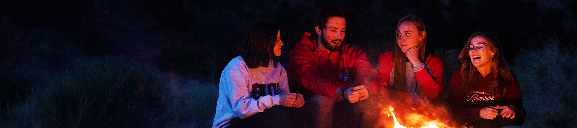 Four young people sit around a campfire at night. 