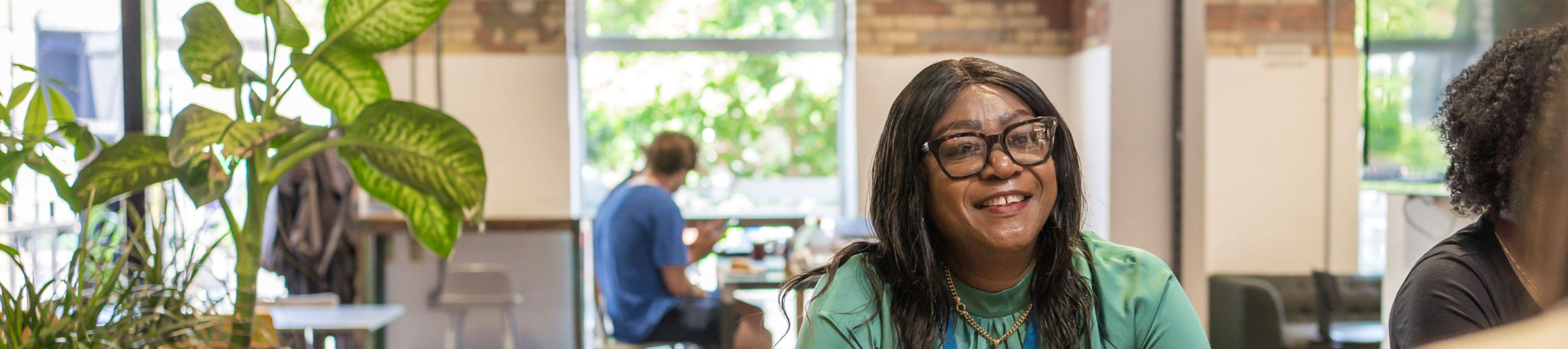 Professional in green top working at a wooden table in a bright co-working café with natural light, brick walls, and indoor plants — modern workspace with collaborative atmosphere.