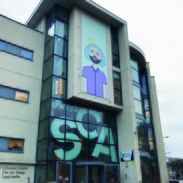 Image of Swansea College of Art Dynevor Building, a beige brick building with glass front with SCA in white writing with graphic image of boy in purple t-shirt