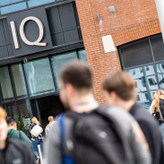 Mae myfyrwyr yn siarad y tu allan y fynedfa i adeilad brics modern lle mae’r llythrennau arian IQ yn sefyll allan o baneli du uwchben y drysau llithro gwydr.