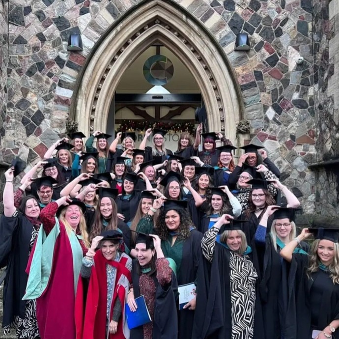 Mae tua deugain o raddedigion Blynyddoedd Cynnar mewn dillad academaidd yn sefyll o flaen drws carreg bwaog Gothig Fictoraidd, gwên, a dal eu capiau academaidd.