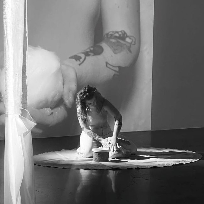 In a monochrome photo, a woman with long hair kneels on a circular piece of cloth and kneads an object with her hands; a video is projected onto the wall behind her. 