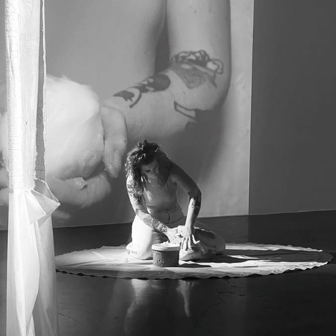 In a monochrome photo, a woman with long hair kneels on a circular piece of cloth and kneads an object with her hands; a video is projected onto the wall behind her.