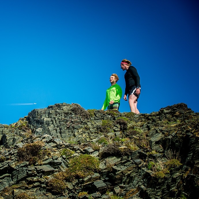 Two young men in running gear stand on an outcrop of rock below a bright blue sky.