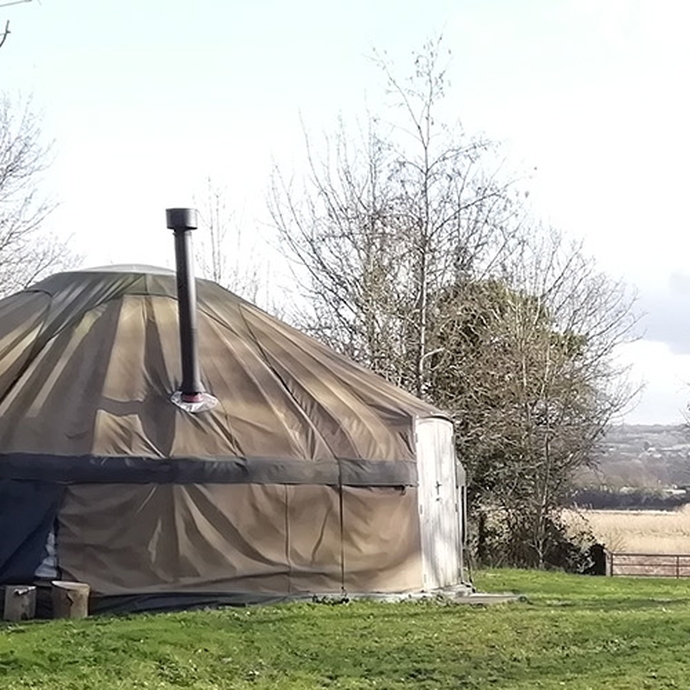 A large canvas yurt in a small field; the yurt has a metal chimney; there is a seating area outside with logs and picnic benches; river meadows and the Towy itself are visible in the background. 