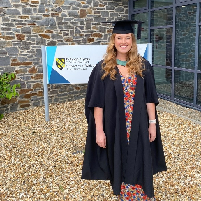 Sarah Bolton standing in her cap and gown in front of the University's sign  on graduation day 