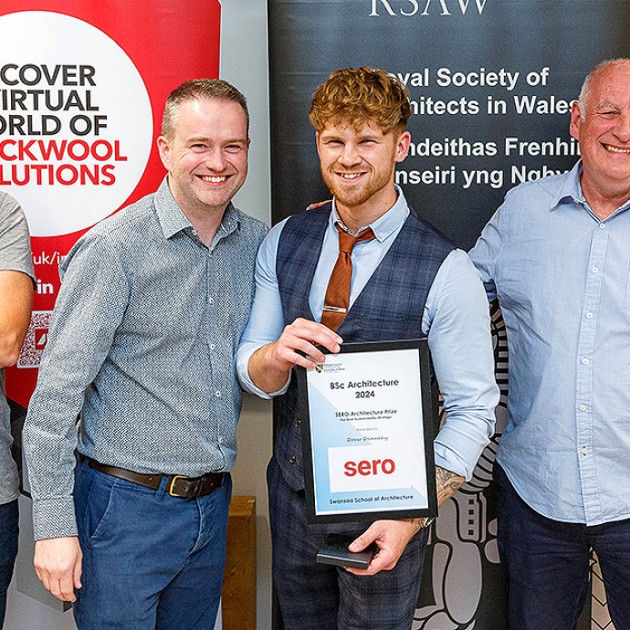 Drew Dennehy, a young man with blond hair wearing a shirt and tie under a dress waistcoast, holds his BSc Architecture and smiles broadly; he's standing between three older men, all smiling, including Architecture programme director Ian Standen.