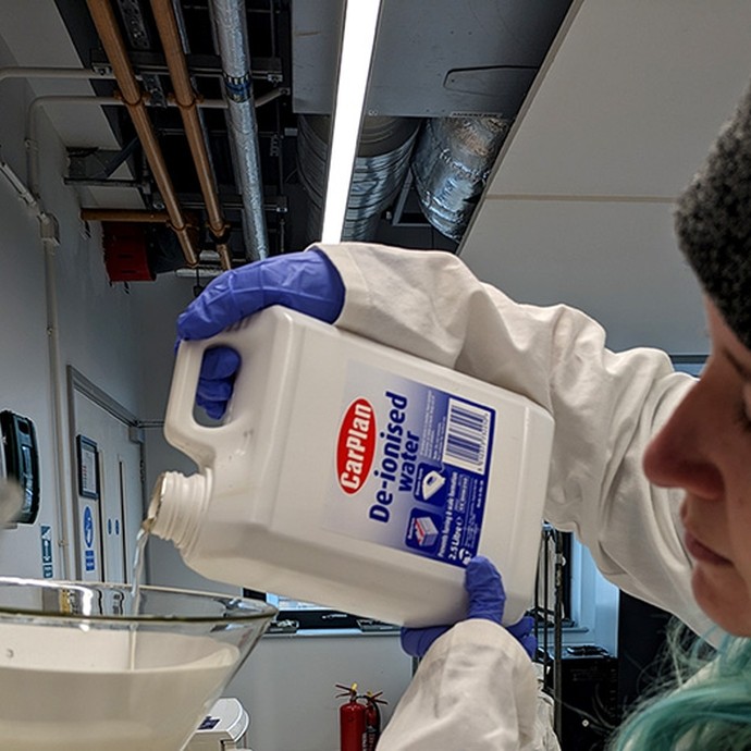 Wearing a woollen hat, a white labcoat and rubber gloves, Rebecca Macfarlane pours liquid from a tub labelled De-ionised Water into a plastic funnel.