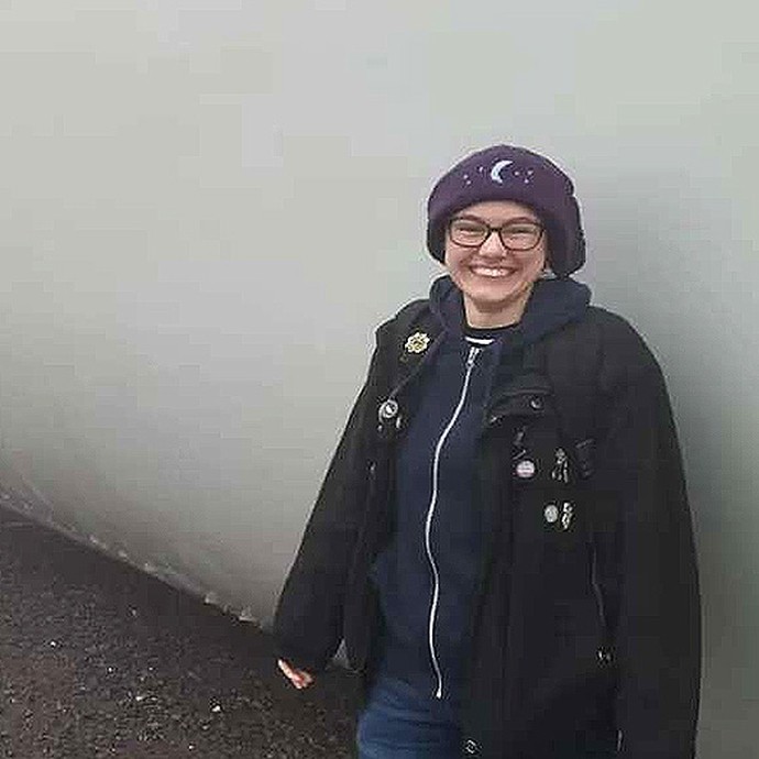 Wearing a purple hat with a crescent moon in its centre, Rowan Jack Moses stands smiling in front of a large, long shape that might be the hull of a ship. 