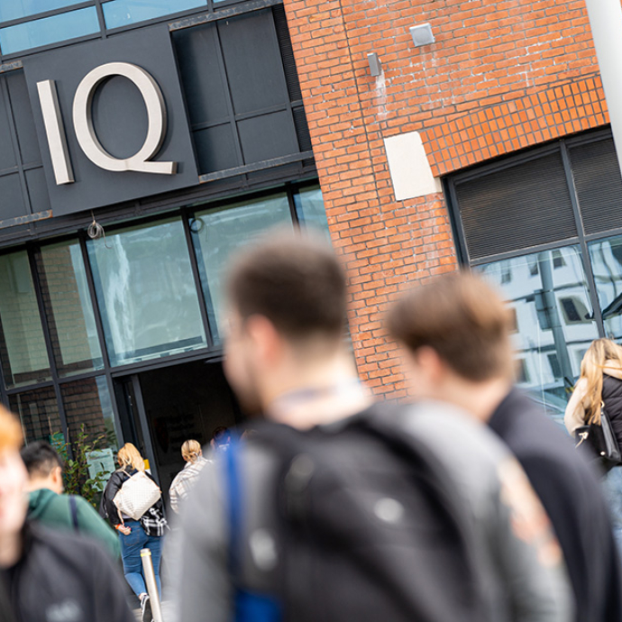 Students chat outside the entrance to a modern brick building with the silver letters IQ standing out from black panels above the sliding glass doors. 