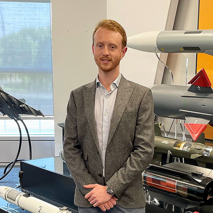 Wearing a grey jacket over an open-necked shirt, Max Hart stands in front of a display of missile casings, including a Sea Skua.