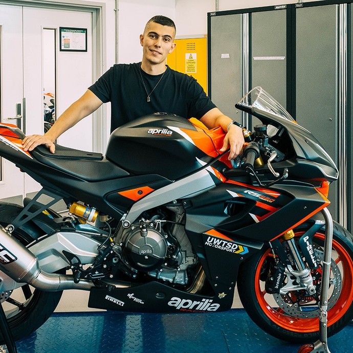 Vinicius Dreher standing and smiling, hands resting on the saddle and fuel tank of an Aprilia racing motorbike. 