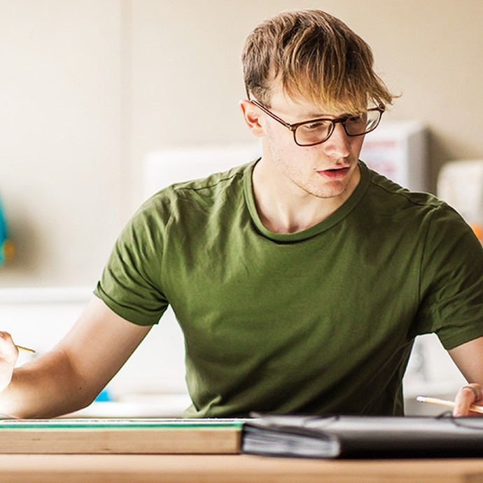 A young man with blond hair, glasses and a green t-shirt works at a desk with a brush in one-hand, cross-referencing a detail. 