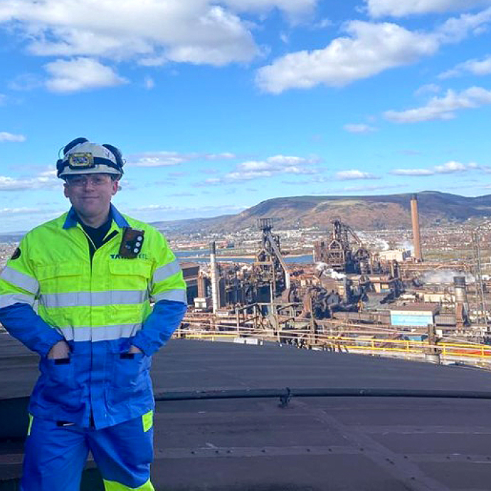 Wearing a hard hat and fluorescent overalls, Daniel Richards stands on the roof of a cooling tower; the Port Talbot steelworks and hills behind Briton Ferry form the background. 