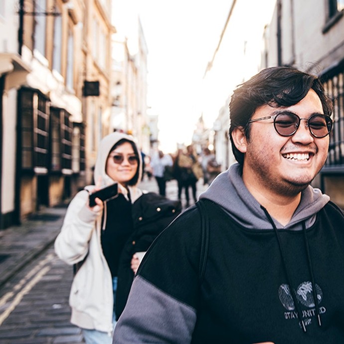 A young man wearing round shaded glasses grins as he walks down a cobbled street. 