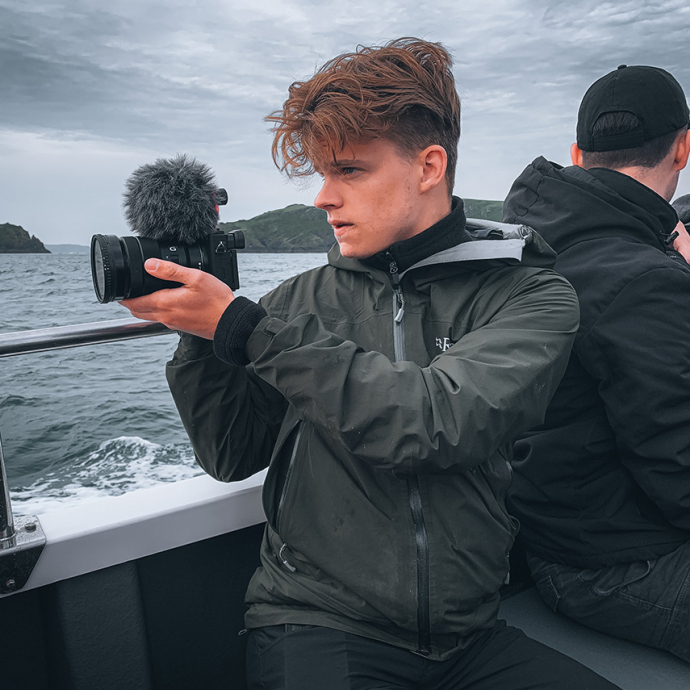 On a grey day, Joshua Knight sits near the stern of a boat and turns towards the sea, holding up a camera with a microphone attachment.