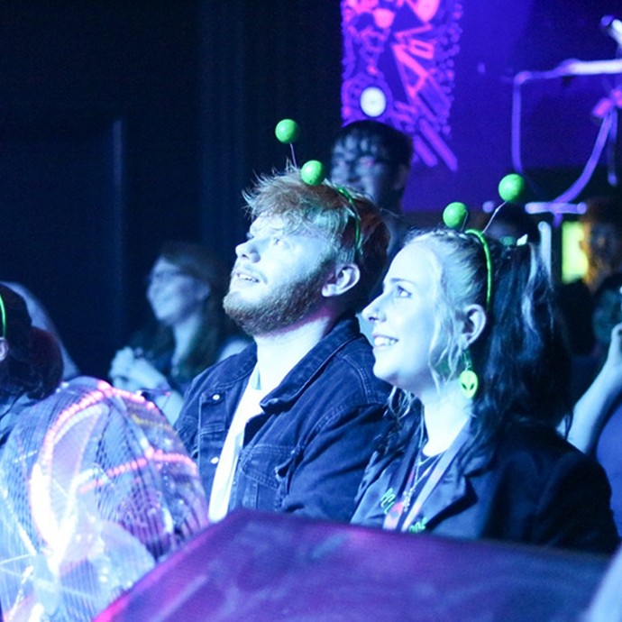 In a busy room lit with purple light, a young man and woman wearing green antennae head-boppers look up towards something off-camera.