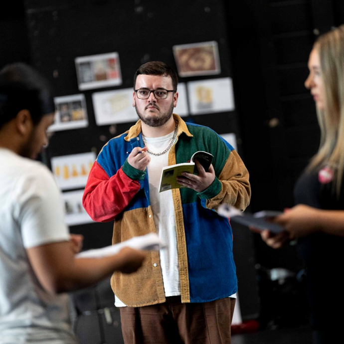 William Kingshott stands in a rehearsal room; he holds a playscript open in one hand and has a pencil in the other; his eyes are focused on another student. 