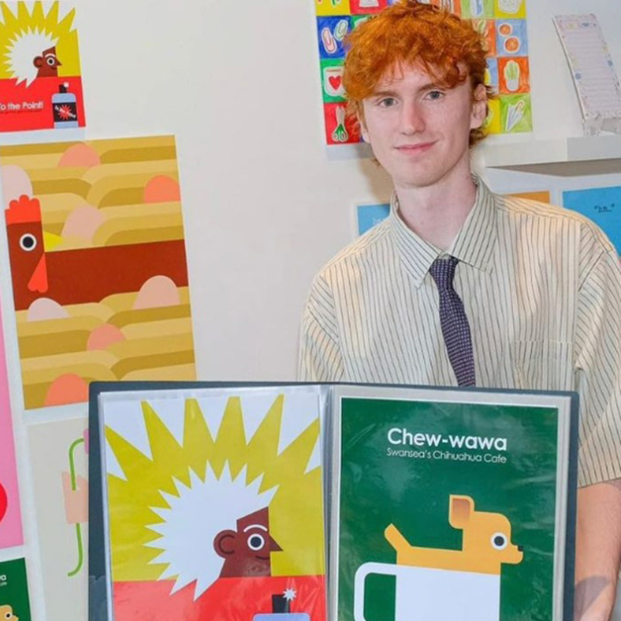 Evan Elias smiles slightly towards the camera as he stands by an exhibition of his illustrations.