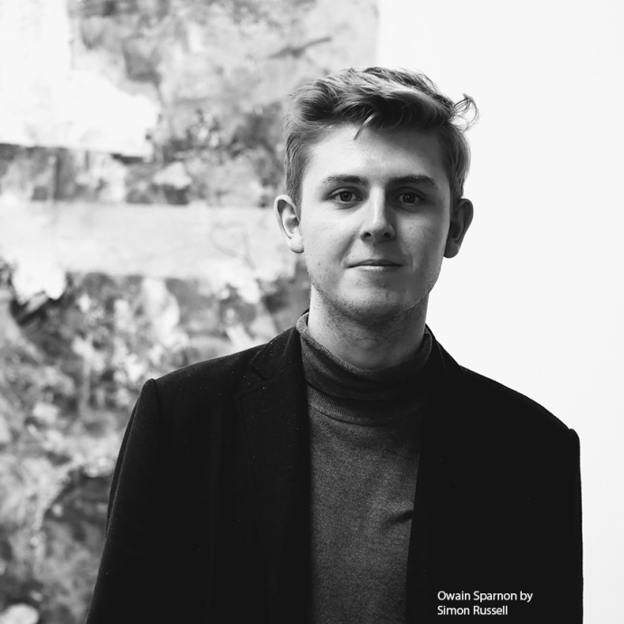 A black-and-white photo of Owain Sparnon wearing a black coat over a polo-neck; he is standing in front of an abstract, textured artwork.