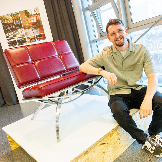A smiling student, seated next to the chair, with his designs on a board in the background.