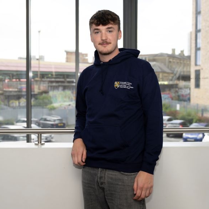 A male student standing in front of a large window, wearing a university branded hoodie.