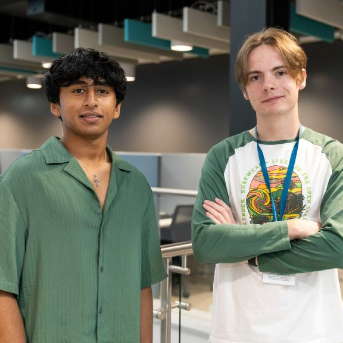 Two male students, one of whom has his arm folded, standing against a glass handrail.