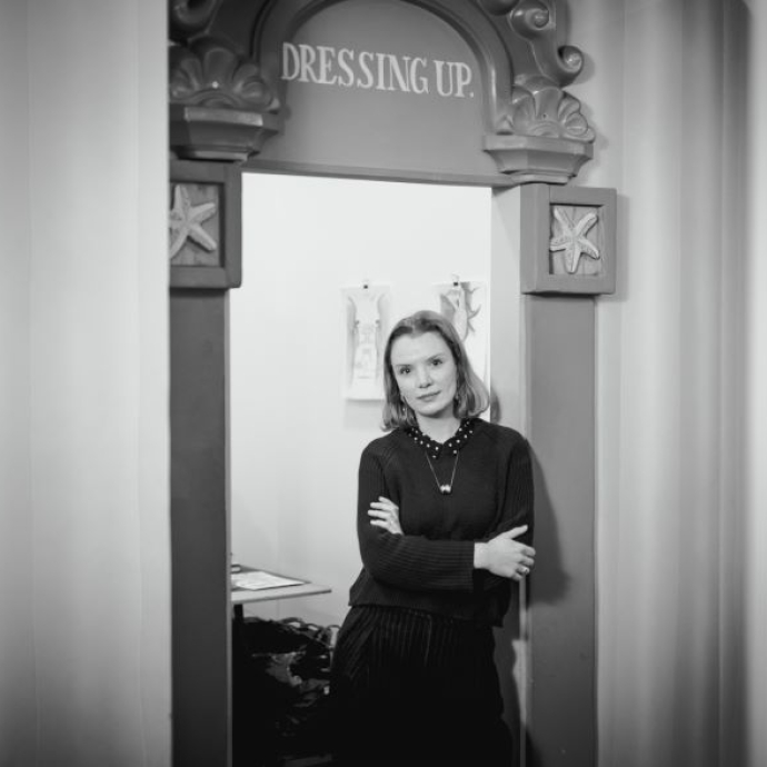 A black and white portrait image of the artist standing in a doorway.