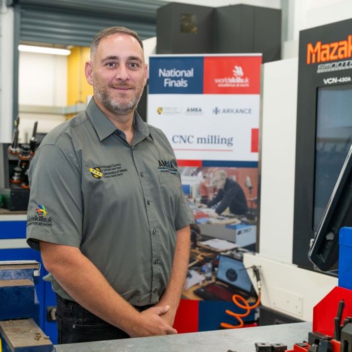 A man standing with his arms folded, wearing a branded top, in a workshop environment.