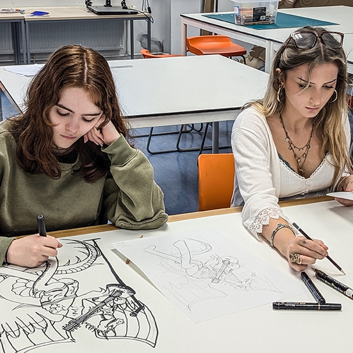 Two college students looking down at their illustration work, sitting at a large white table in a workshop.