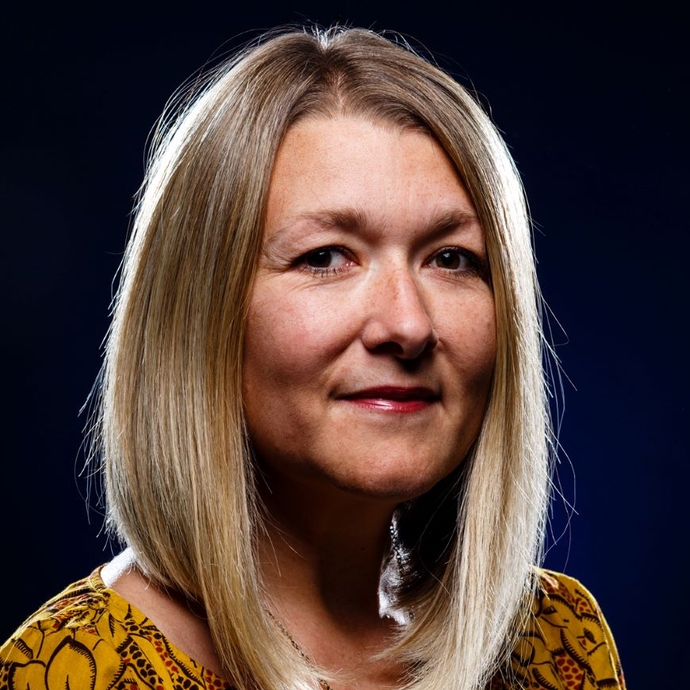 A smiling lady dressed in a patterned top, looking at the camera, against a black backdrop.