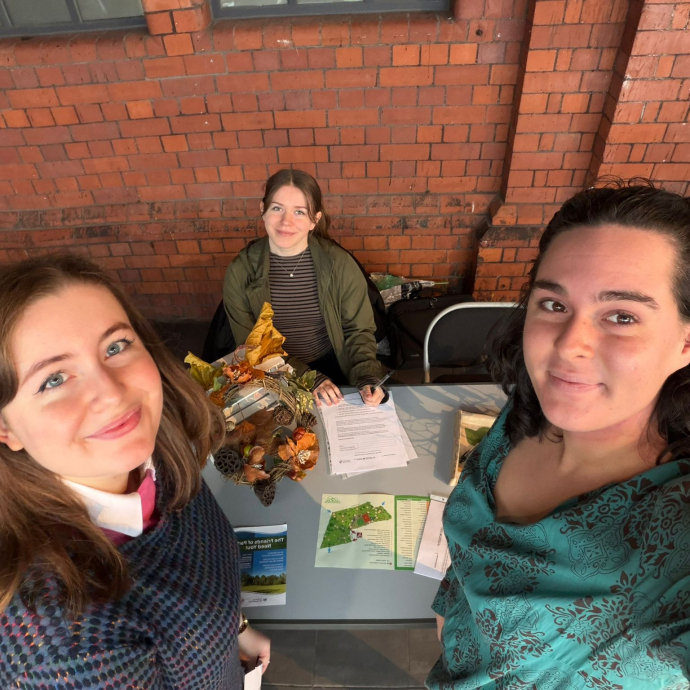 Three students, two of which are standing and one is seated, next to a table covered with leaflets and pens.