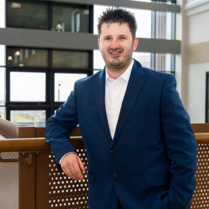 A graduate standing in front of a glass bannister on the top floor of a new building.