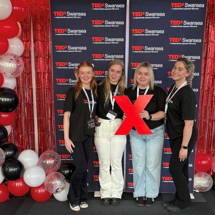 Four students standing between a red, white and black balloon arch in front of a red and black branded background.