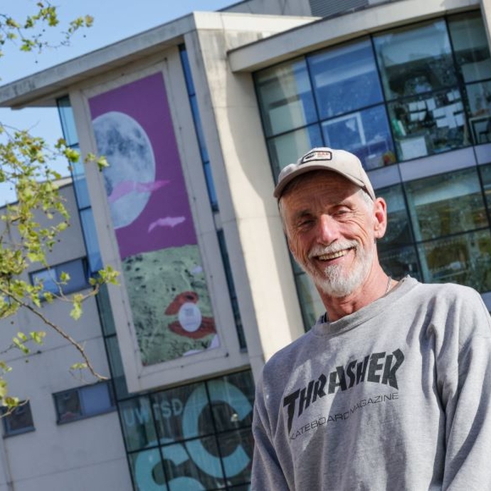 A man in a baseball hat and grey sweatshirt smiling and standing outside a glass-fronted building with a giant canvas at the top.