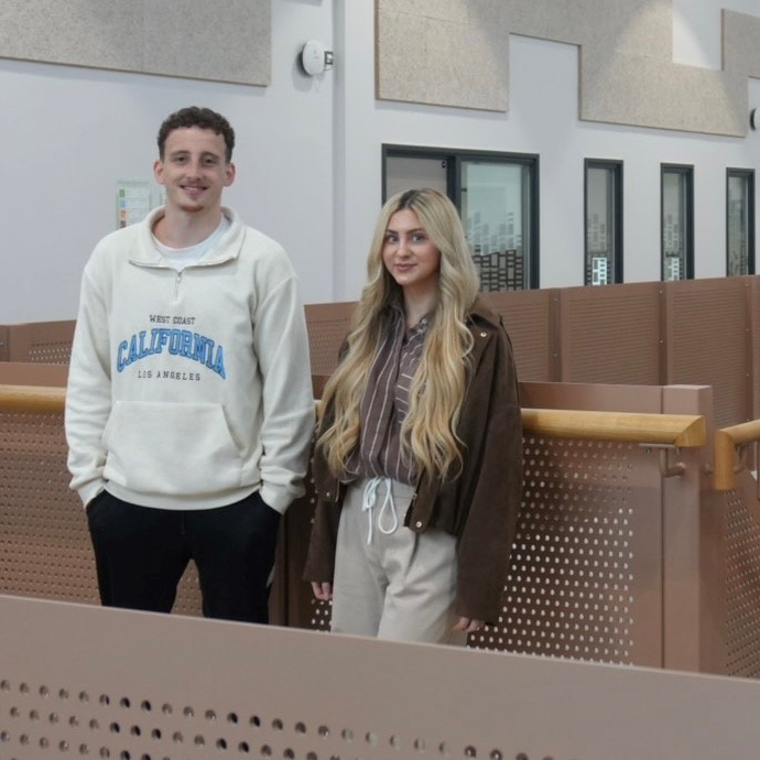 Two students, one dressed in a white hoodie and dark trousers, and the other in a dark top and light trousers, stand smiling in front of a wooden and glass bannister.