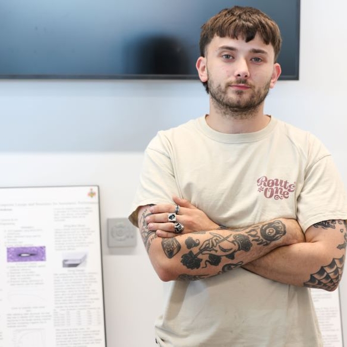 A student in a light top, standing with his arms folded in front of an exhibition board.