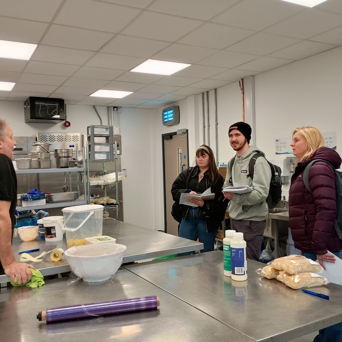 Three students in a catering kitchen at the university talking to a chef