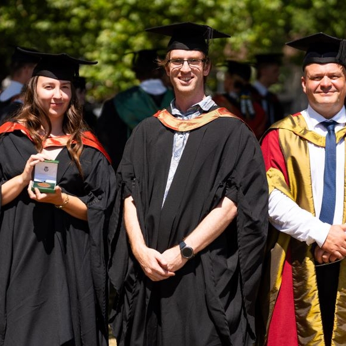 A garduate and two lecturers smiling and standing proudly in their caps and gowns.
