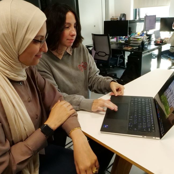 Two University staff sat at a white desk looking at technology.