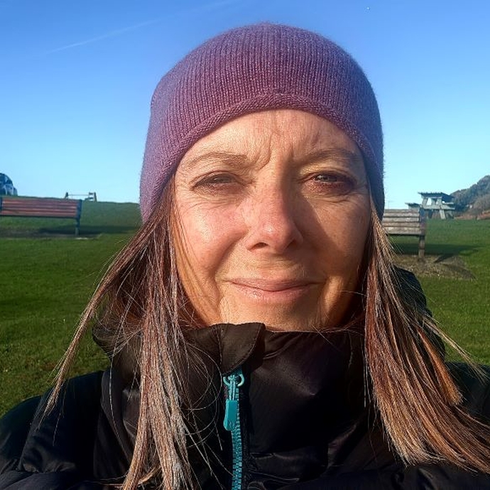 A lady with long hair, wearing a hat, standing outside, with green grass behind and blue sky above.